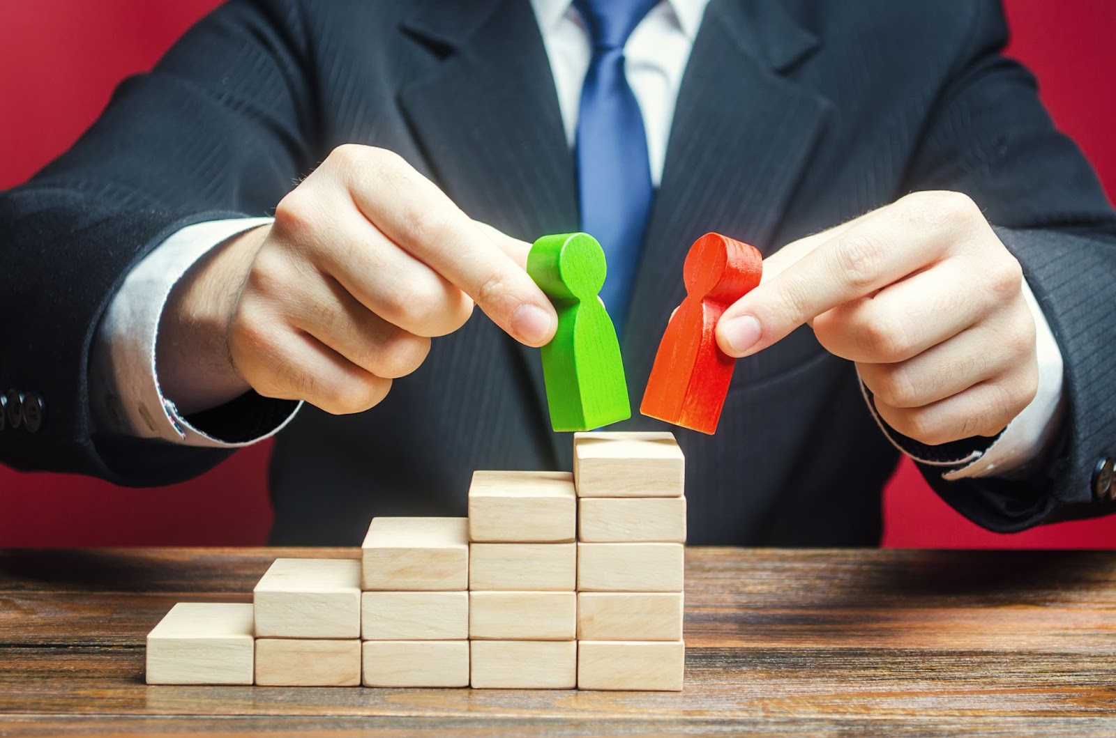 A close-up of a businessman in a black suit, face hidden, holding red and green faceless wooden dolls on stacks of wooden blocks to symbolize change.