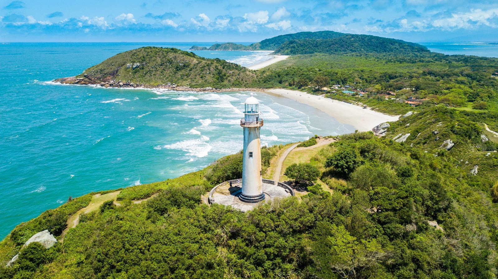 Praia do Farol, na Ilha do Mel, Paraná. O território é quase inteiramente coberto por áreas de Mata Atlântica Verde, que se estendem por montes e encostas. Em uma das elevações, há um farol branco. O mar é azul claro e banha as faixas de areia clara