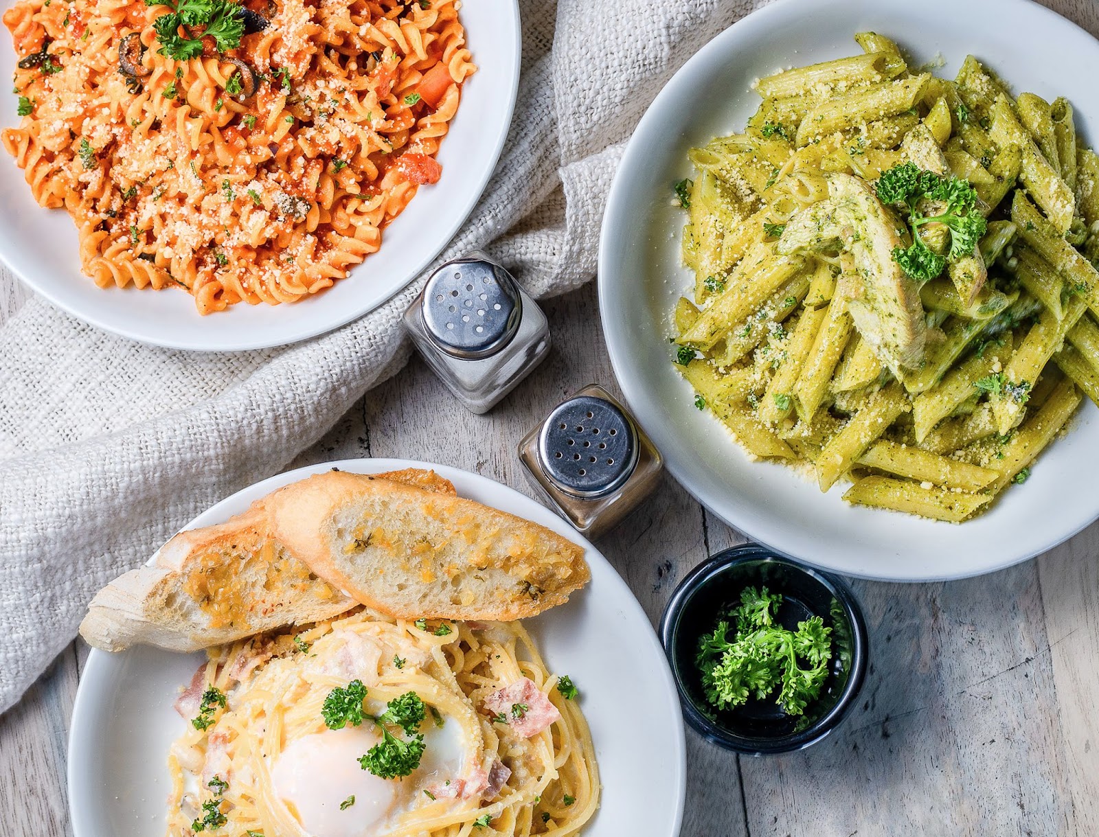 a top-down view of three plates of different types of pasta at a kelowna italian restaurant, with a salt and pepper shaker and small bowl of parsley in the centre of the table