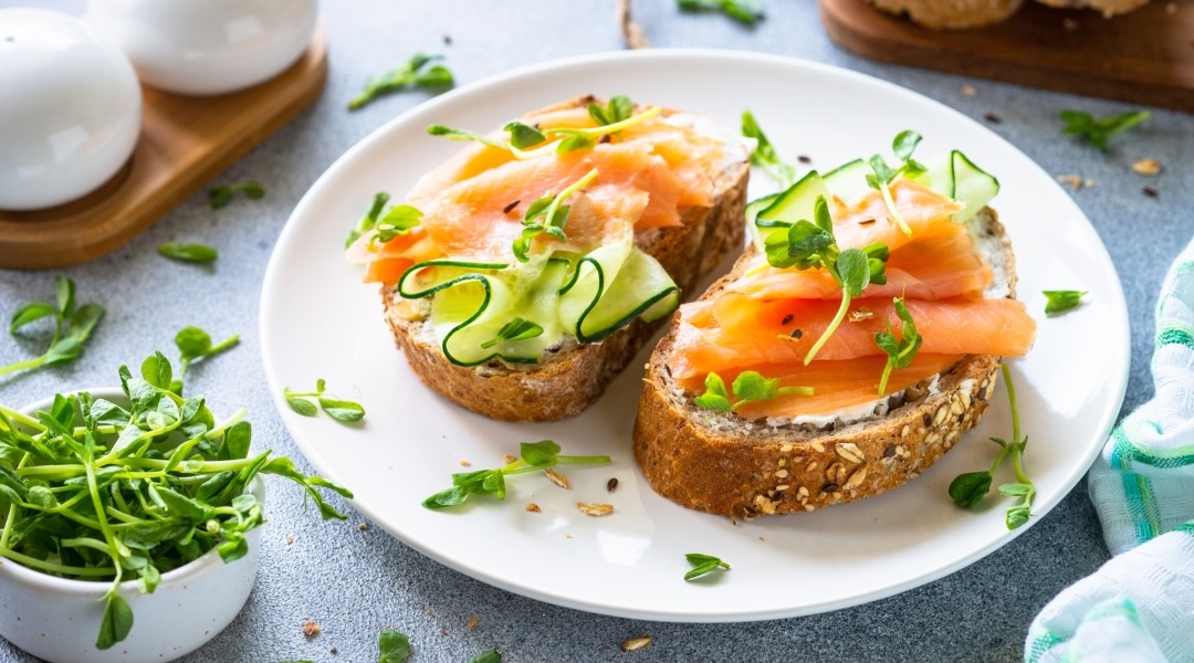 Two slices of whole wheat toast topped with smoked salmon and cucumber slices, served on a white plate placed on a grey countertop.