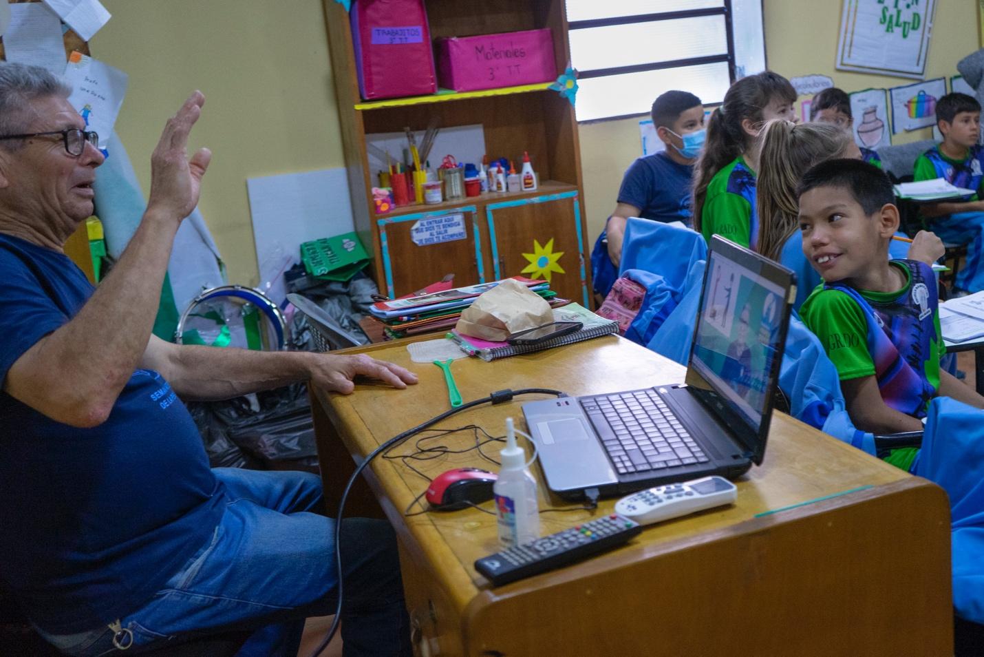 En la clase, el docente tiene una computadora en frente y los niños y niñas participan de la clase