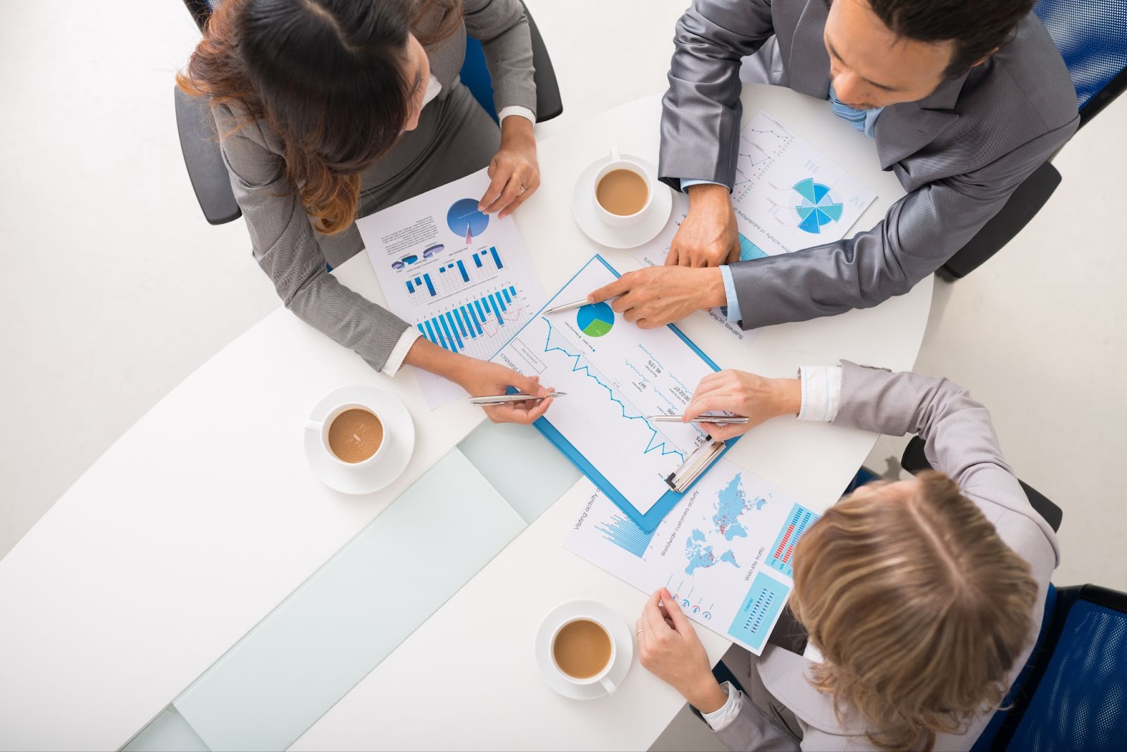 A top shot of three entrepreneurs dressed in gray business suits engaged in a discussion about risk mitigation plans. Assorted graphs and coffee are on the table, highlighting a focused business meeting.