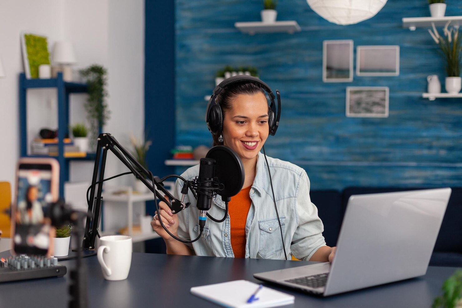 Mulher branca por volta de 25 anos, grava podcast com microfone e fone de ouvindo em frente a um computador portátil. Veste uma camisa laranja embaixo de um casaco jeans