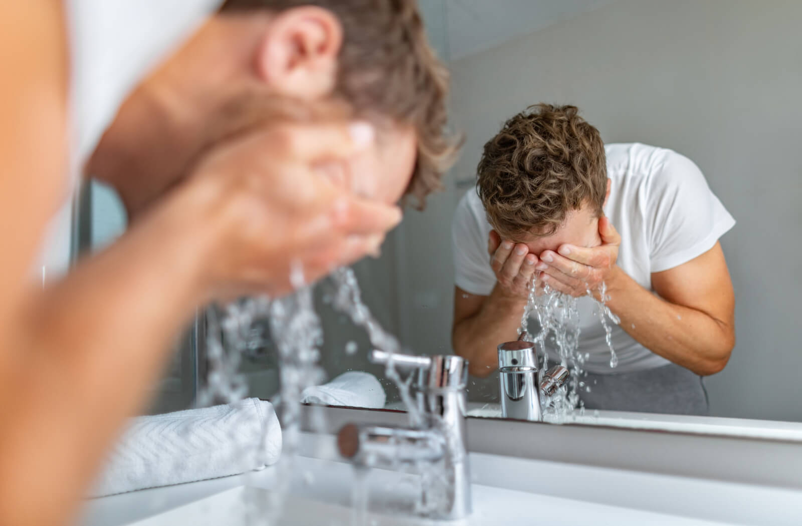 A man washing his face with water in front of a bathroom mirror.
