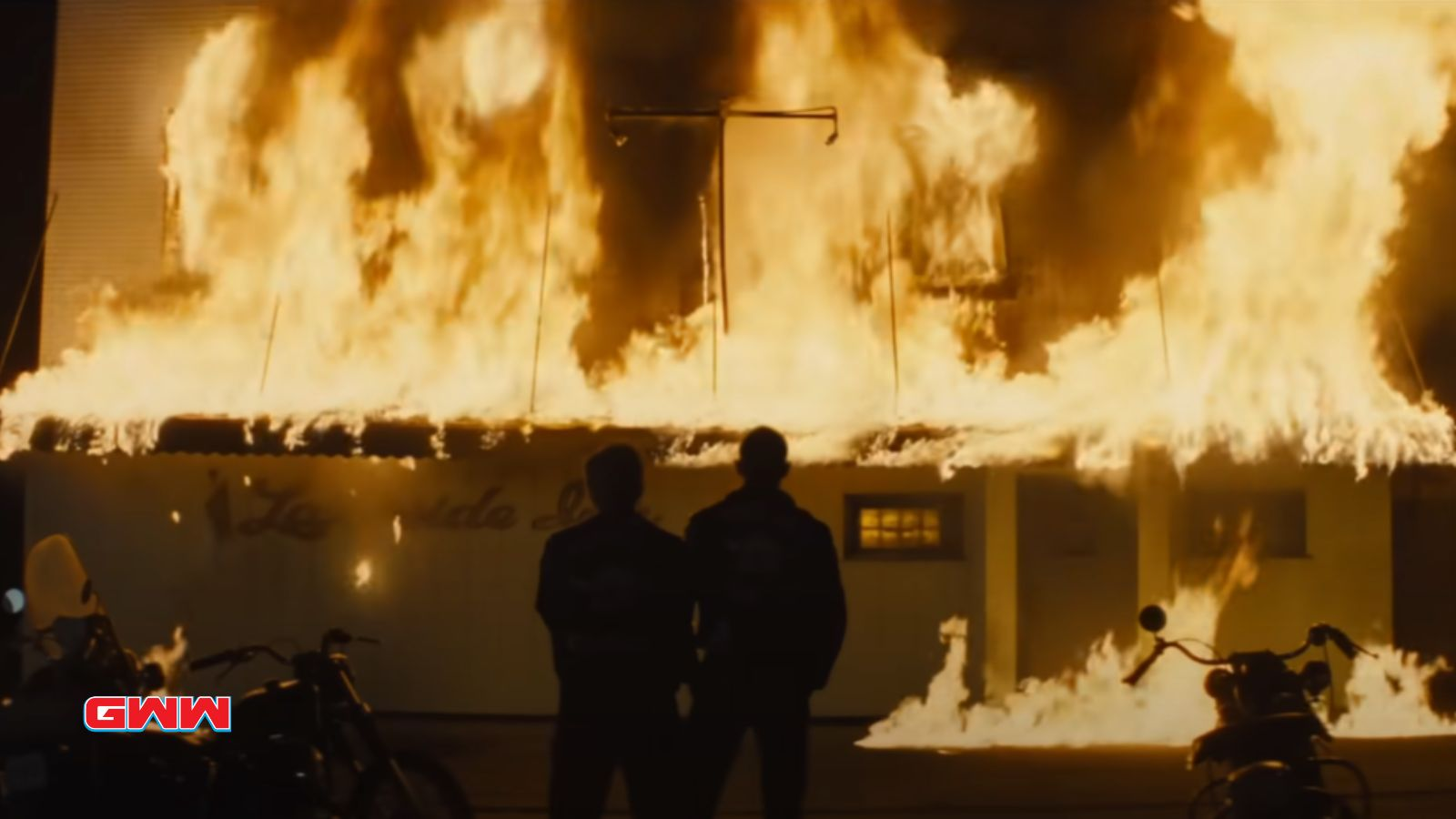 Building engulfed in flames at night, silhouettes of two people watching.