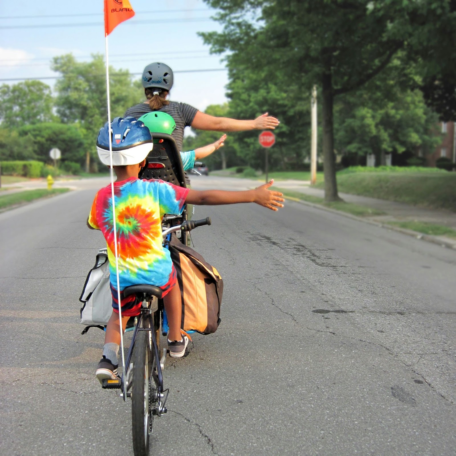 A family riding down the middle of a street as seen from the back. A woman is pedalling the bike with a kid in the bike seat behind her and an extended bike connected with a kid in a tie-dyed shirt in the back. 