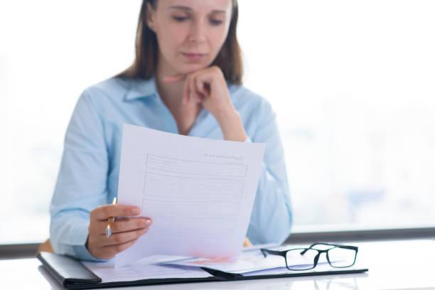 Serious woman reading and examining document Serious woman reading and examining document. Entrepreneur sitting at desk and working. Paperwork and recruitment concept. Front view. employee screening and background check stock pictures, royalty-free photos & images