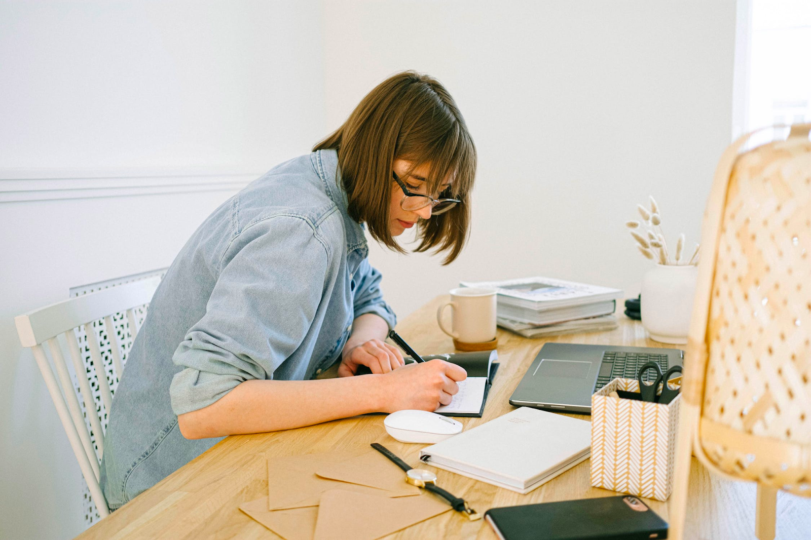 A woman wearing glasses and a denim shirt is sitting at a desk writing in a notebook, this image was added in the blog post Your Website Needs a Blog 