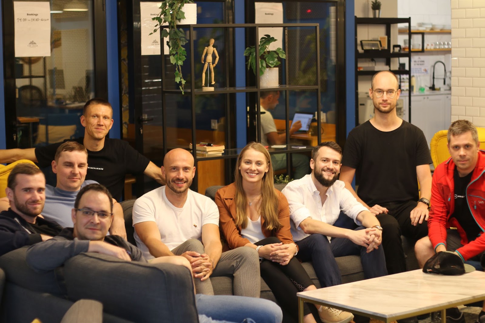 Nine members of the primehammer family (8 men and a woman) pose for a picture while sitting on a couch around a coffee table in an indoor setting.