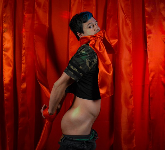 Dakota Wonders posing in a camo crop top against a red curtain with his hands tied behind his back and his mouth gagged