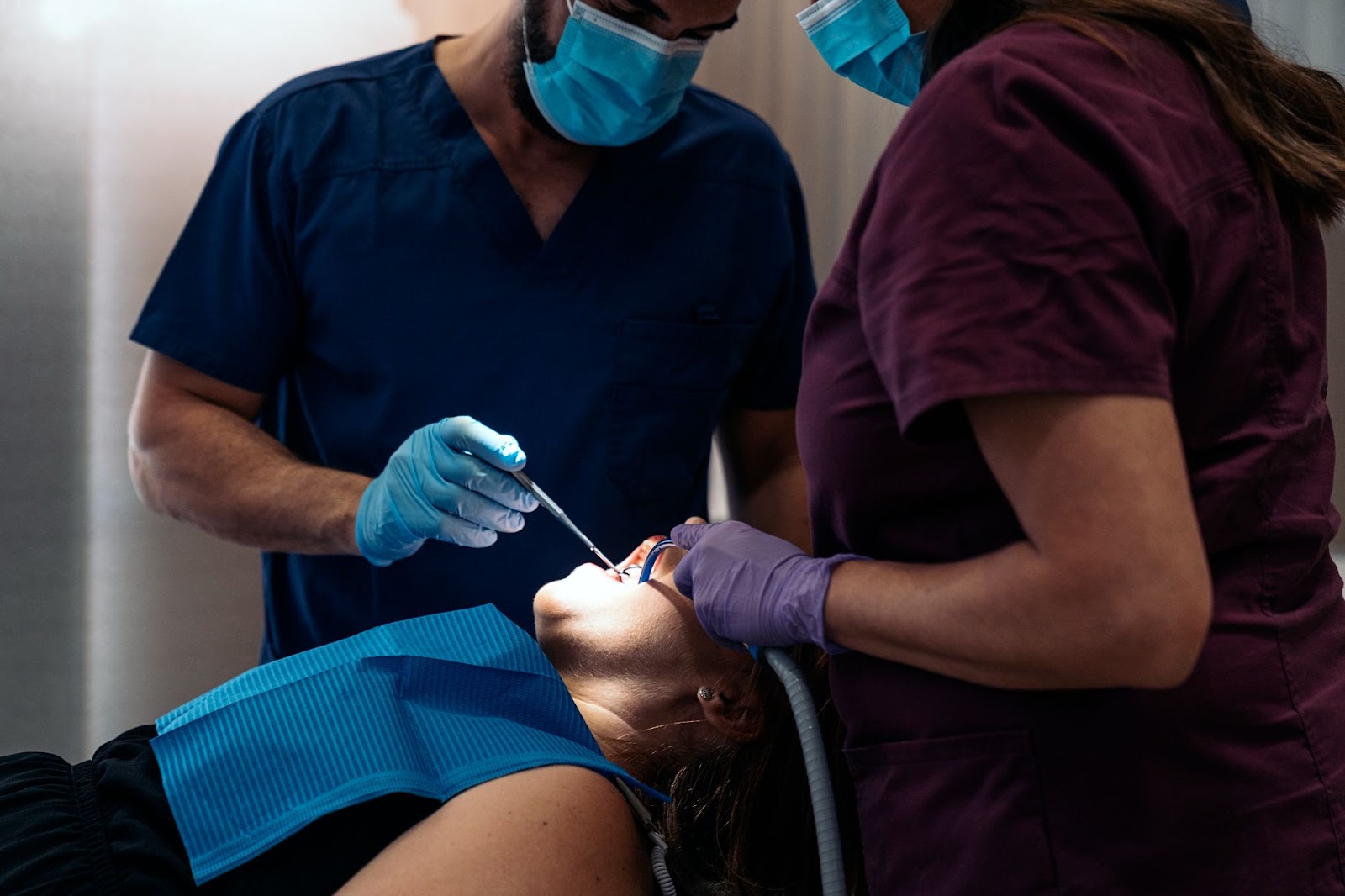 Two dentists in violet and blue scrub suits examine the female patient's teeth. 