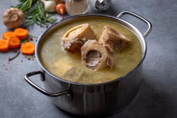 A casserole containing bone broth soup with sliced carrots, whole garlic, and herbs at the background