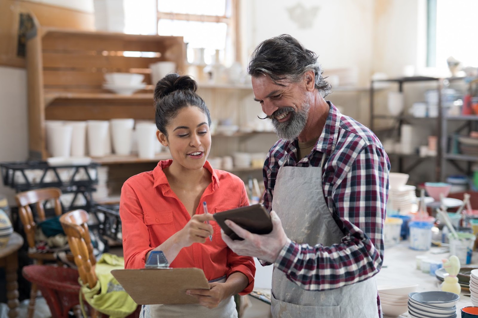 Two business owners discuss business plans over a tablet at their pottery shop, a diverse array of colorful ceramic pieces in the background.