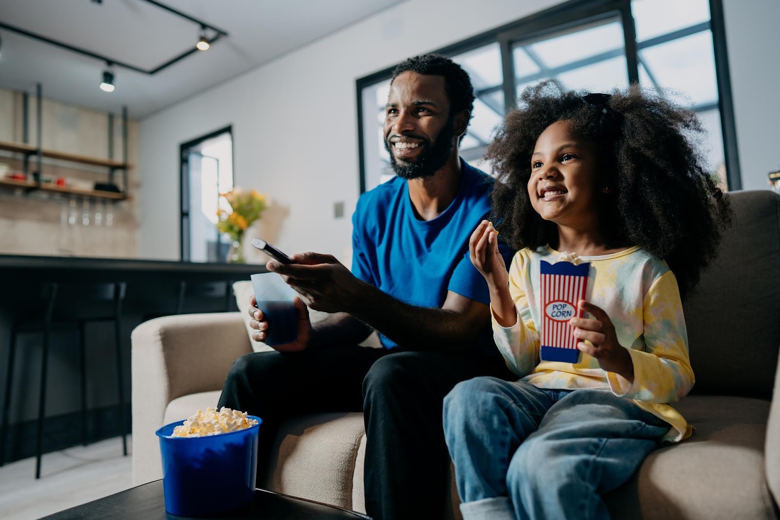 Ein Mann sitzt neben einem kleinen Mädchen auf dem Sofa. Er hält eine Fernbedienung in der Hand, vor ihm auf dem Couchtisch steht ein blauer Eimer mit Popcorn. Das Mädchen hält eine Papiertüte mit Popcorn in der Hand.