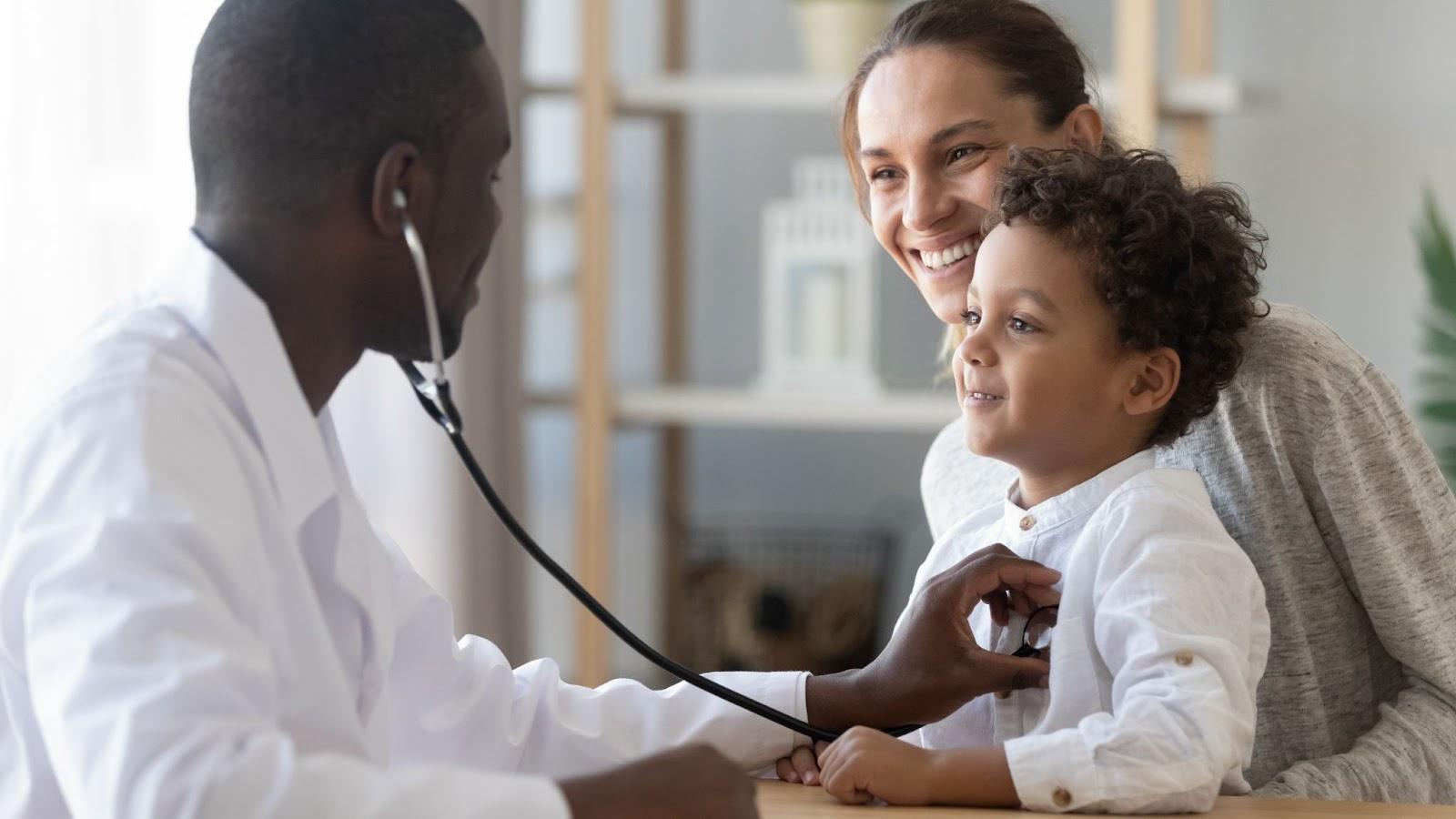 A doctor holding a stethoscope to a child’s chest. The child is sat on his mother’s lap