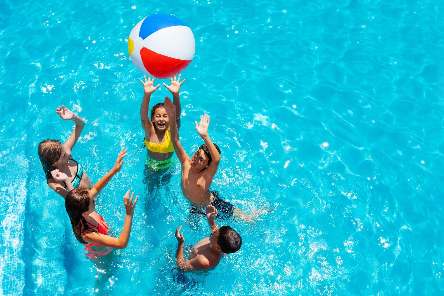 5 kids in a swimming pool jumping up to toss a beach ball