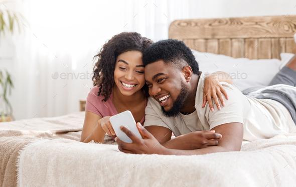 Happy black couple using smartphone on bed Stock Photo by Prostock-studio