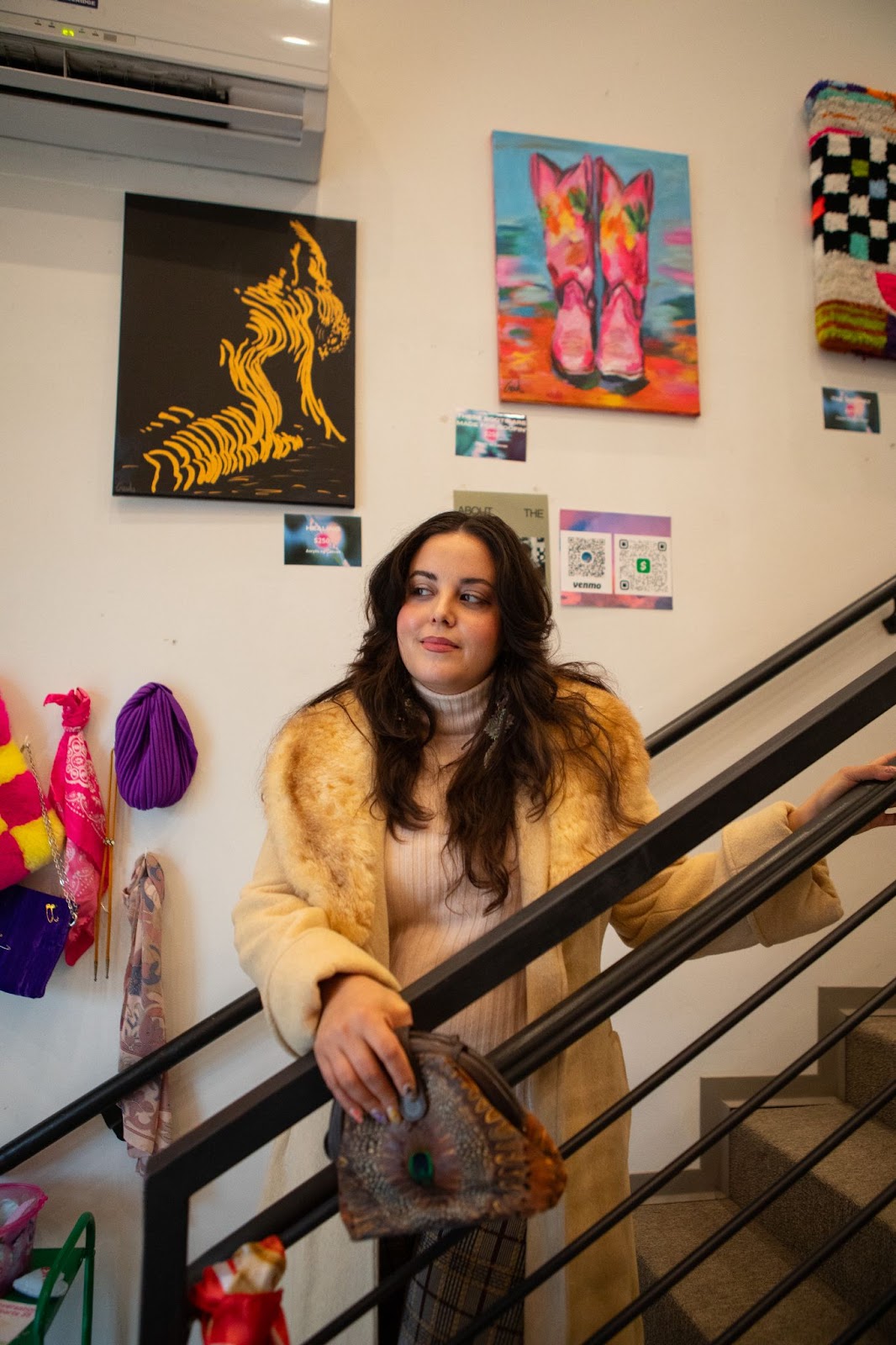 Tesia Zientek standing on a set of stairs in a vintage coat with art pieces displayed behind her on the wall.