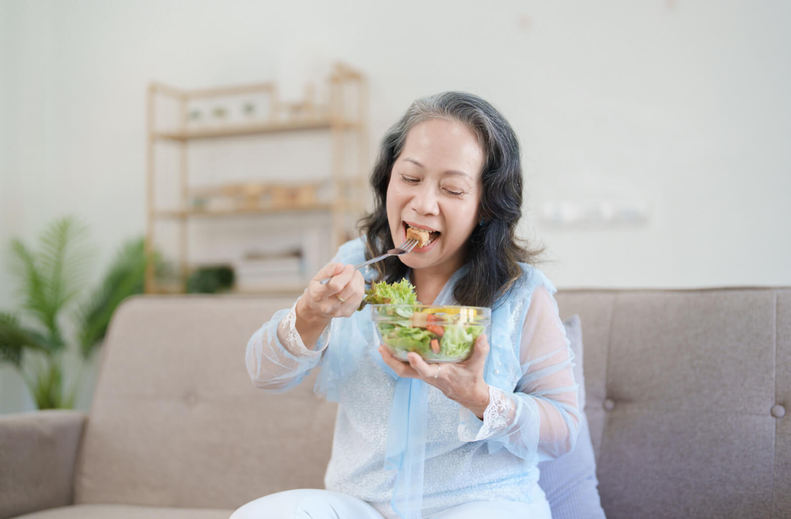 A senior woman eating a bowl of salad in a brightly lit living room.