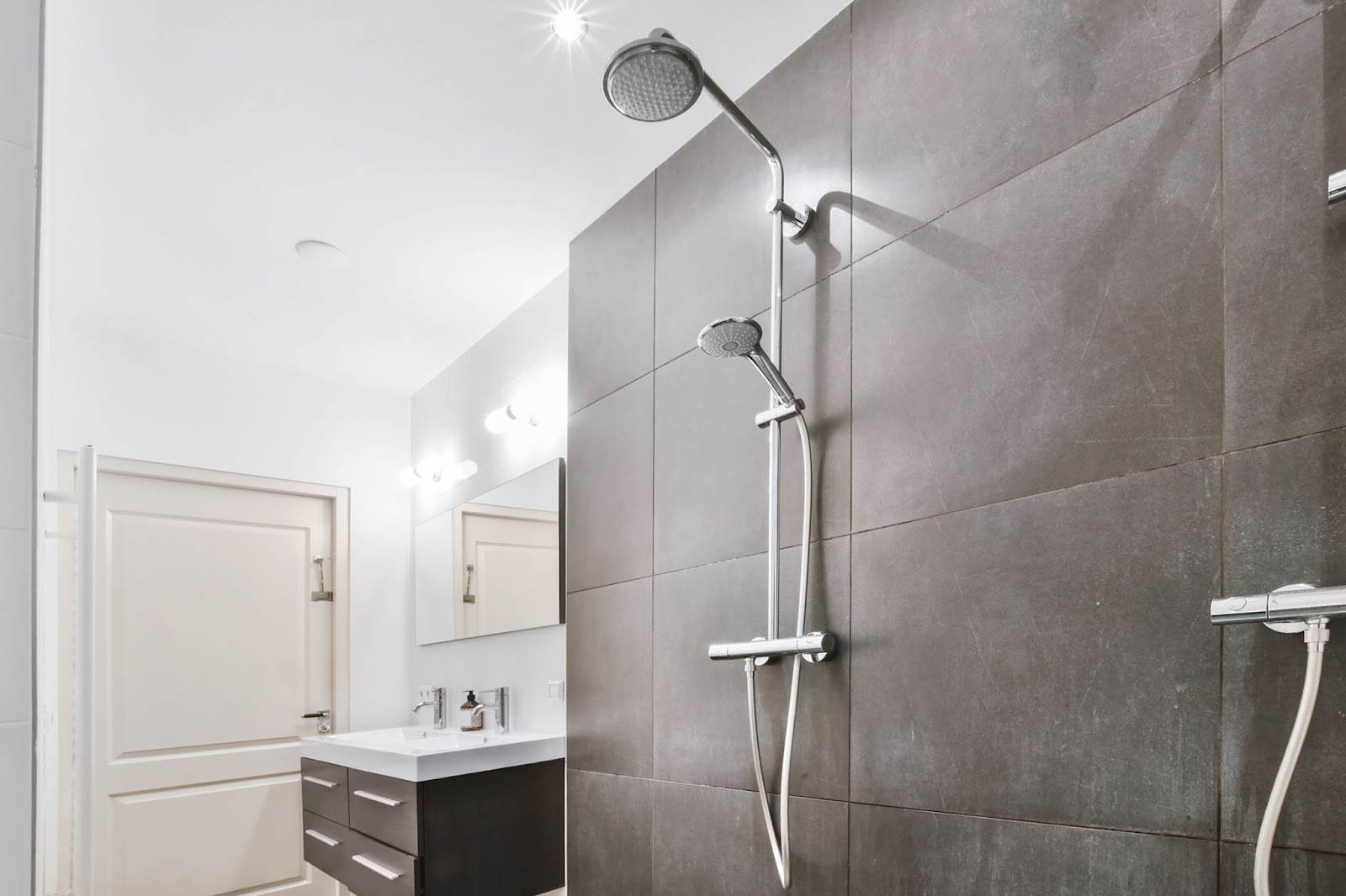 A modern shower stall with gray walls, a sink with gray cabinets, a mirror, and a door in white hues in the background.