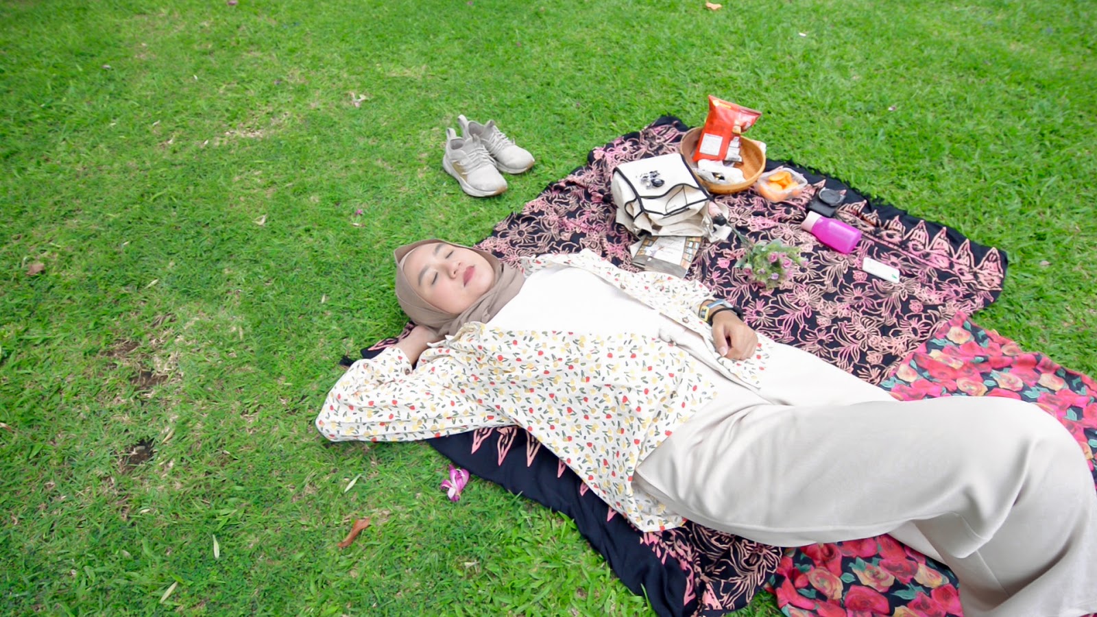 Tips menghadapi cuaca panas, foto seorang perempuan sedang berbaring di atas rumput taman di Jakarta yang rindang, beralaskan kain piknik