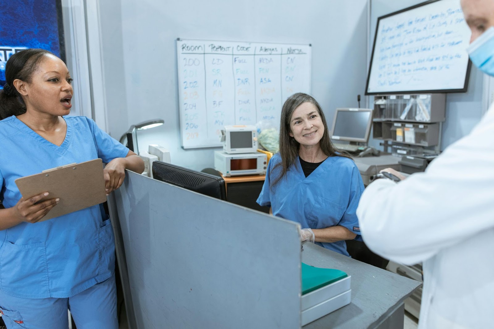 A nurse inputs data on a computer at the nurse's station while consulting with colleagues, with a patient room assignment whiteboard in the background.