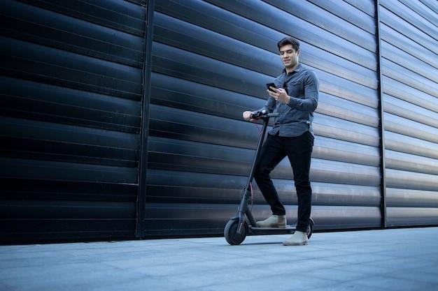 Young elegant man walking with electric scooter and using smart phone