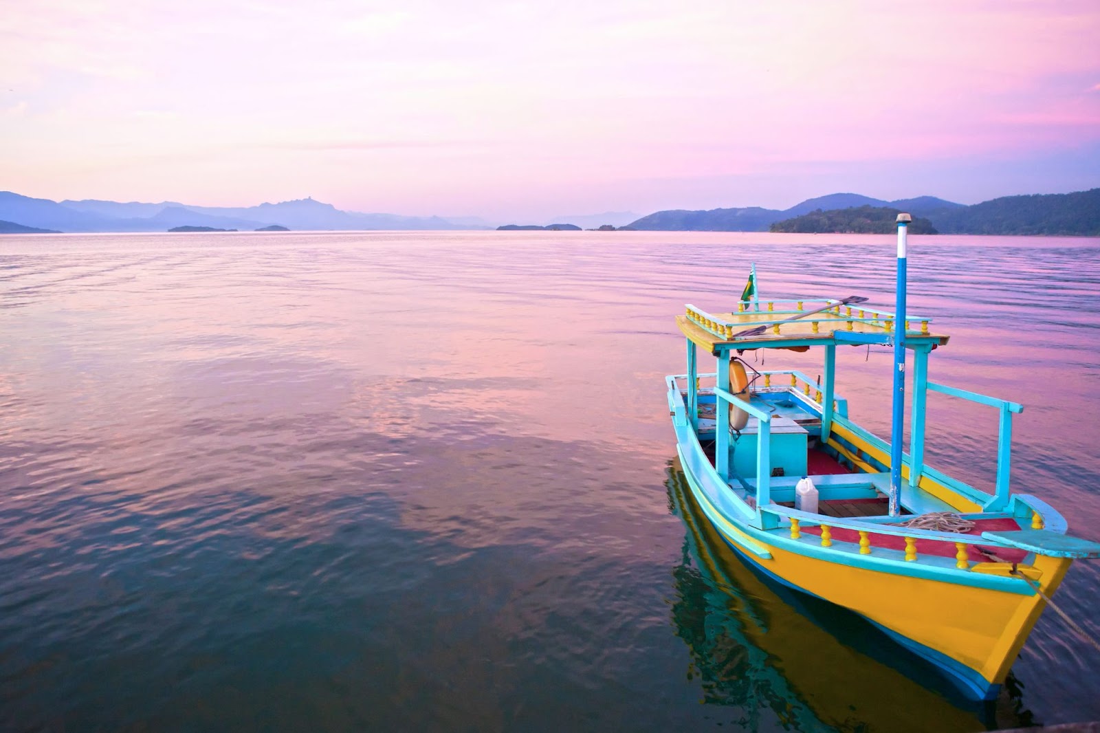 Barco amarelo e azul flutuando no mar sem ondas, em tom rosado refletindo o céu ao entardecer.