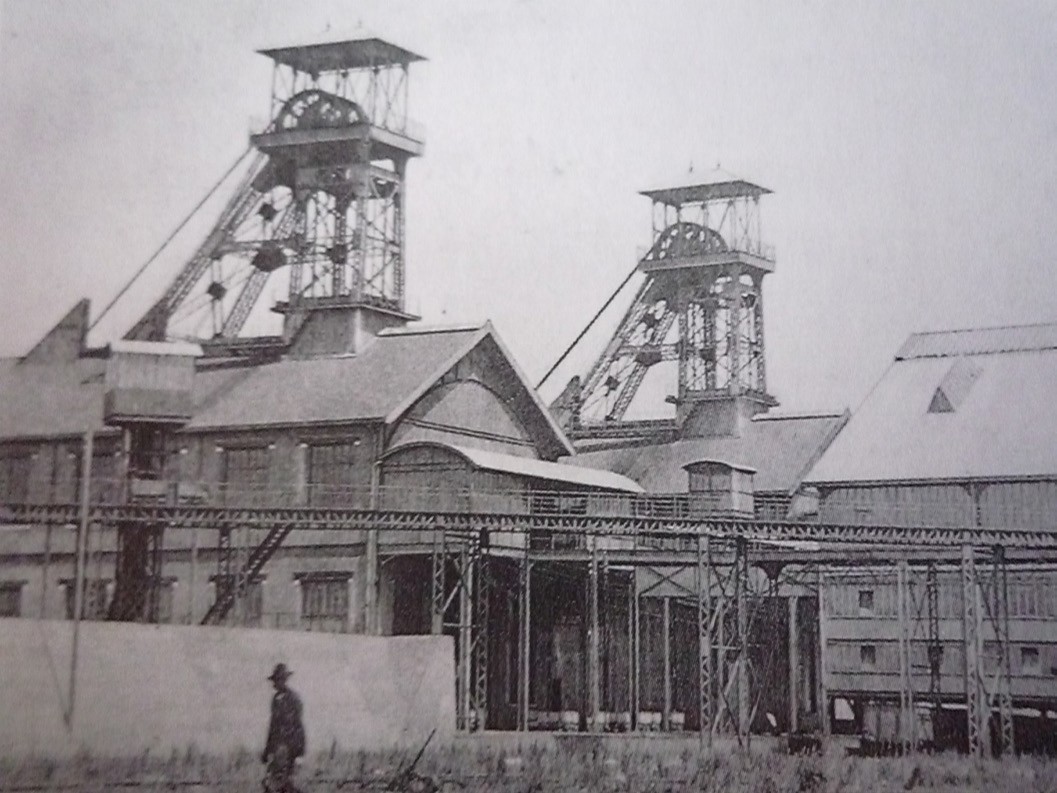 Industrial looking historical buildings with conveyer belts going up to a tower attached to the roof of the buildings. A man in a hat is walking through the foreground. Black and white photograph. 