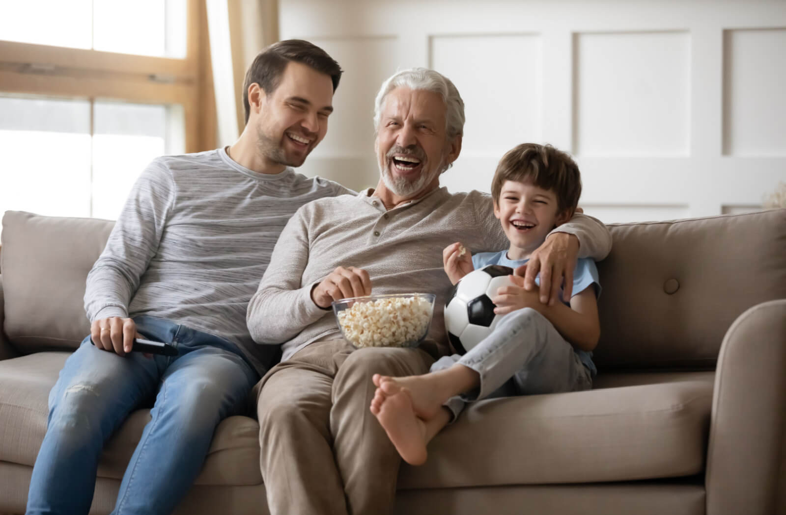 A man visiting his older adult father with his young son; all of them are having a fun time watching TV.