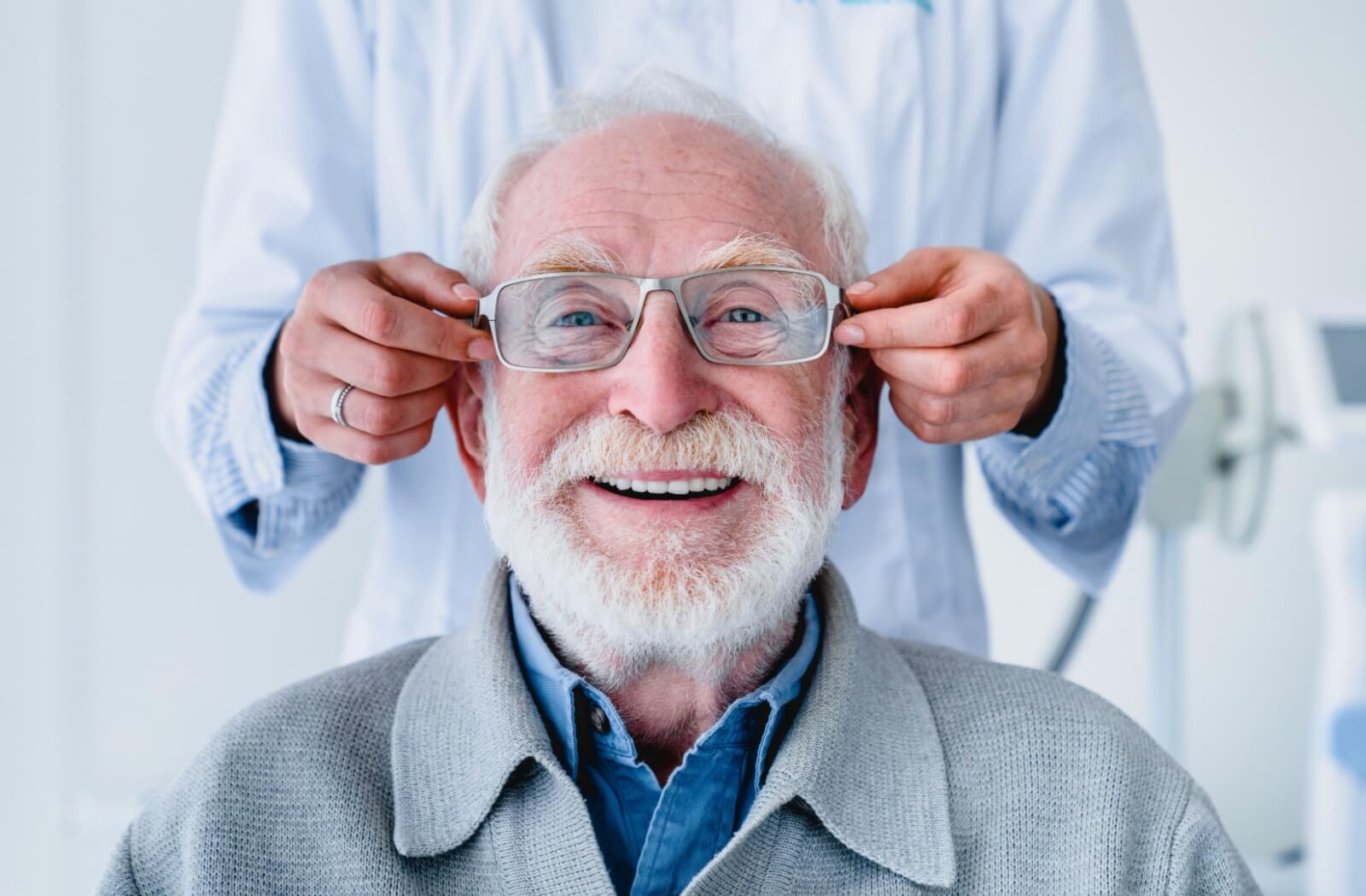 A happy older adult trying on a pair of glasses with the assistance of his eye doctor.
