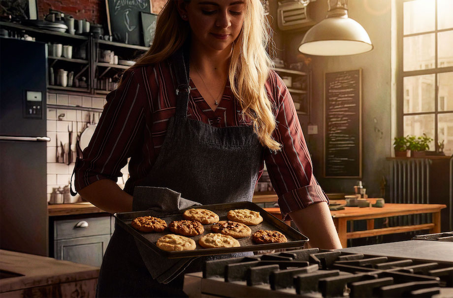 Photo of long haired figure in apron, removing tray of cookie from oven, with AI generated backdrop, by Cincinnati-based food/drink photographer Teri Campbell.