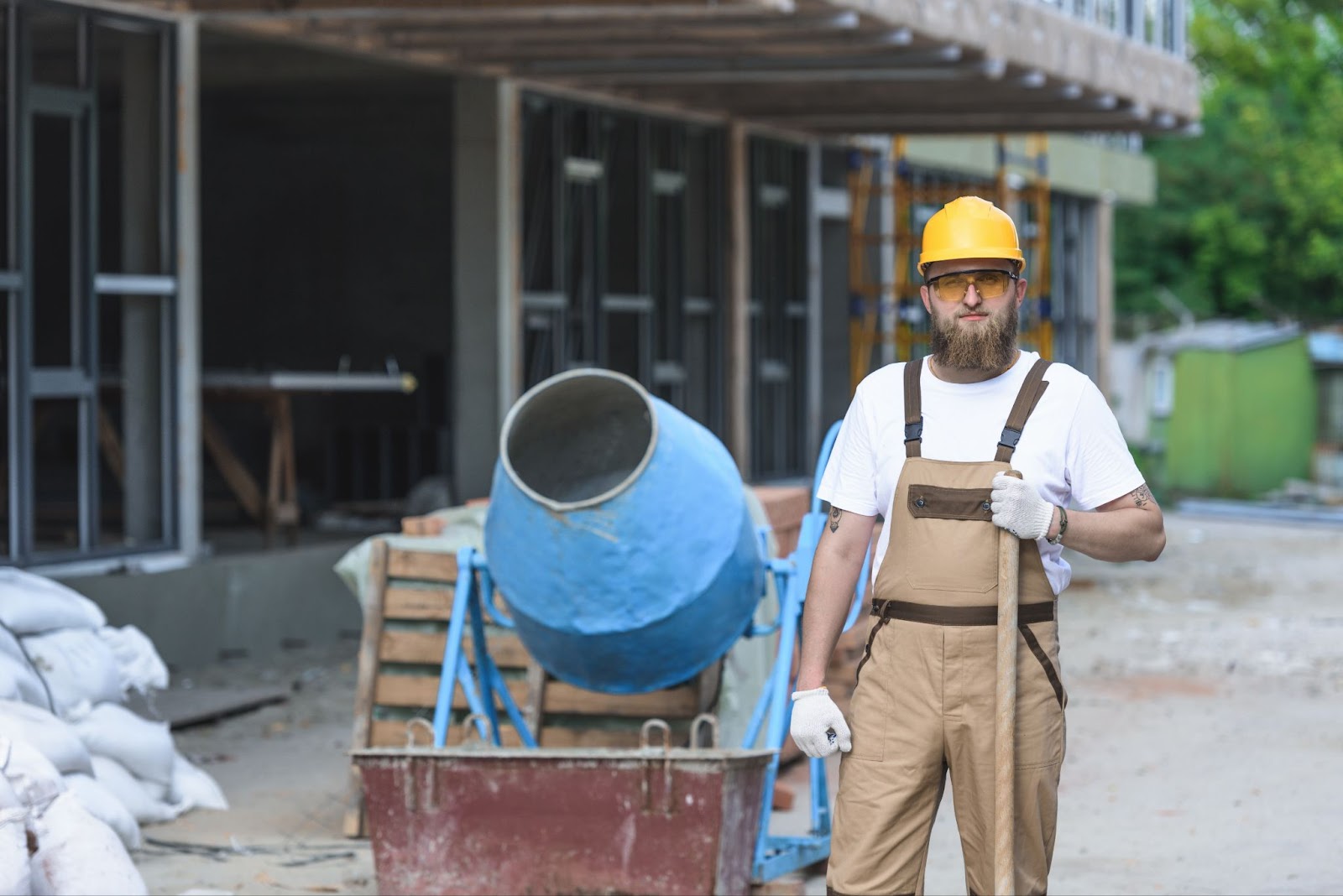 A builder wearing a yellow hat with protective glass and gloves standing near a concrete mixer. 