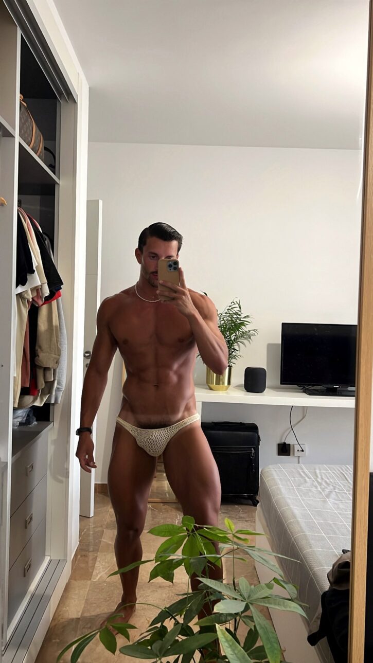 Eric Rmgr posing in front of the mirror in his apartment in a beige crochet bikini showing off his gay bulge and tan