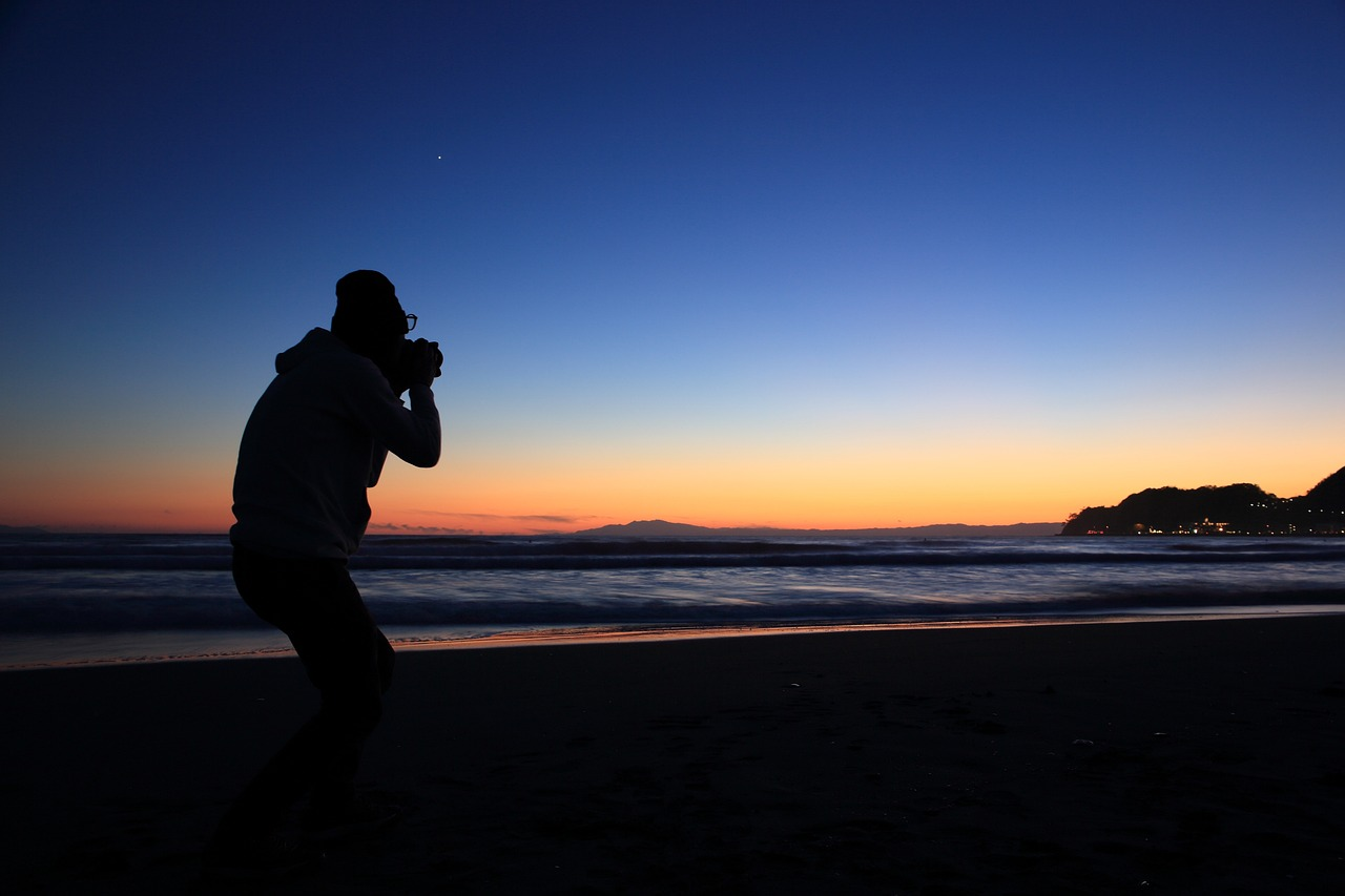 a photographer standing next to the beach against a soft blue and orange sunset and taking a photo