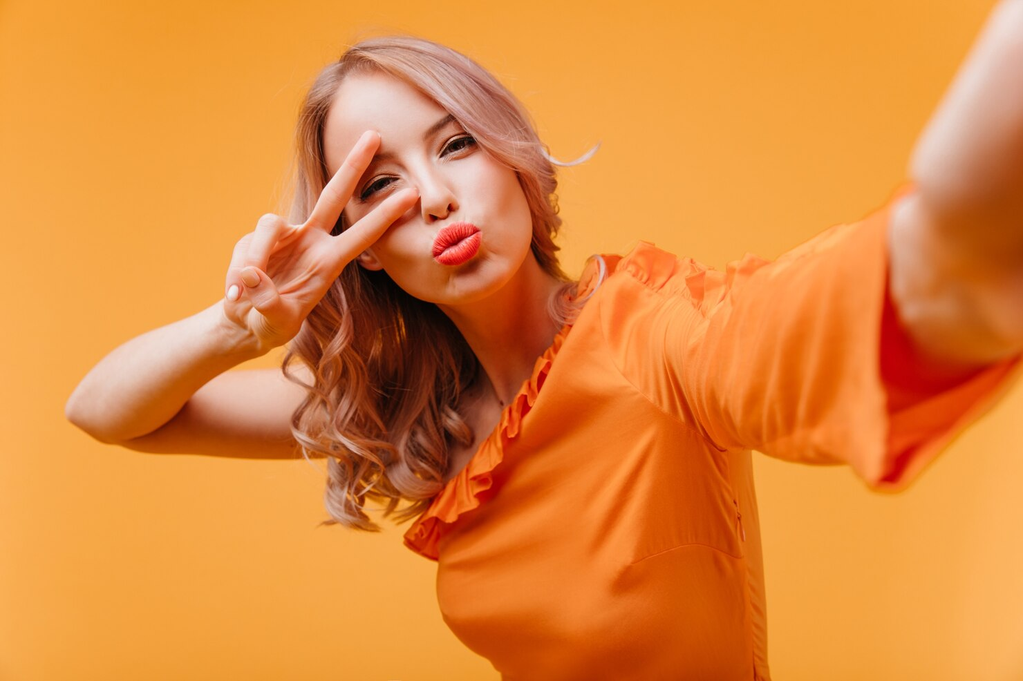 A woman in orange dress making a victory sign and a pout.