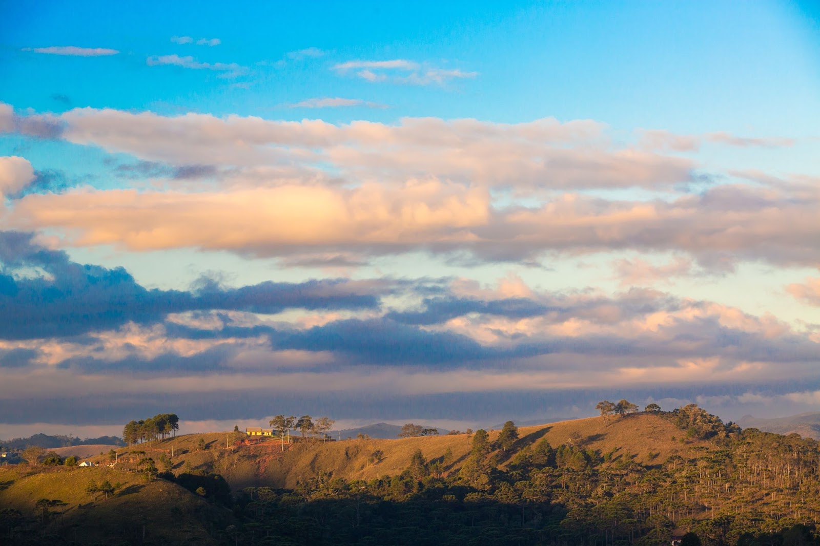 Paisagem da Serra da Mantiqueira em Campos do Jordão. As montanhas cobertas pelo verde da vegetação se estendem até o horizonte. O céu está azul e parcialmente encoberto por nuvens.