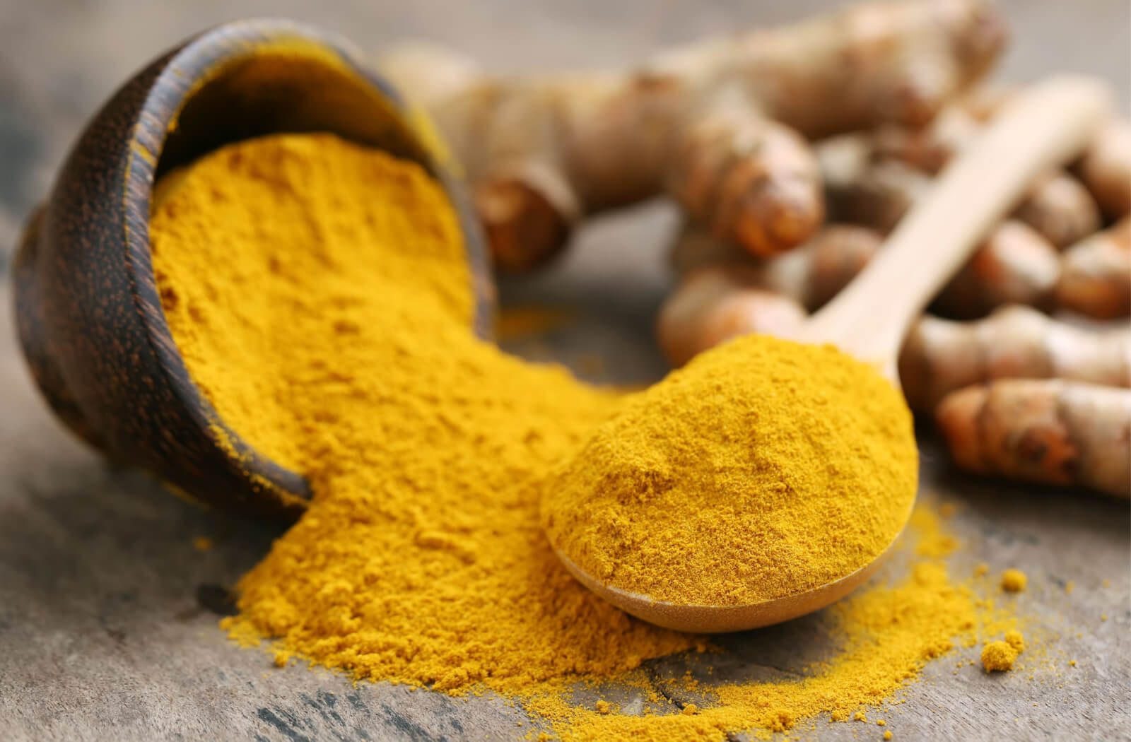 A close-up of a spoonful of turmeric powder beside a bowl of it and turmeric.