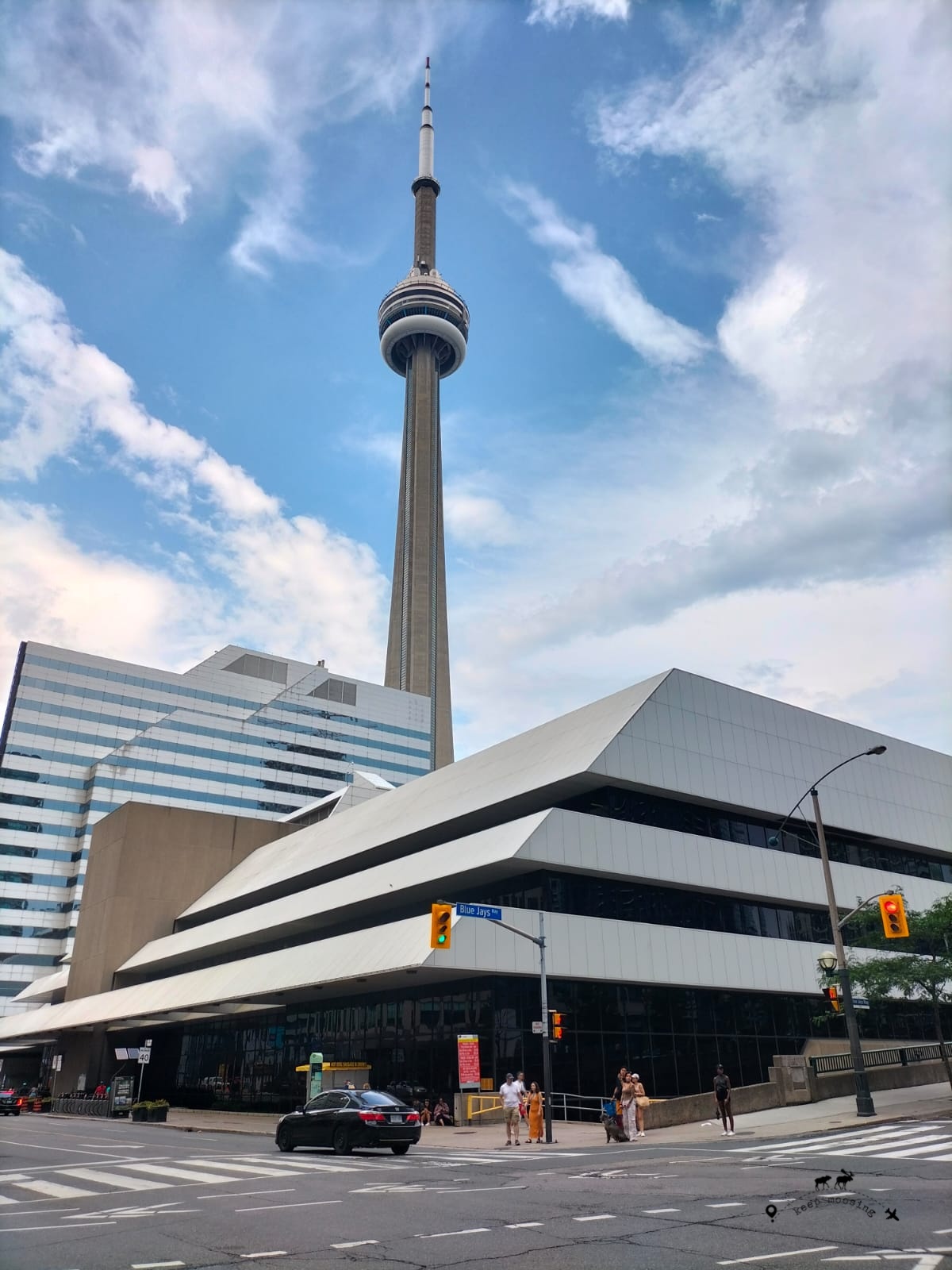 CN Tower rising among buildings in downtown Toronto. In the foreground, a large road intersection and a building with a white sloping roof.