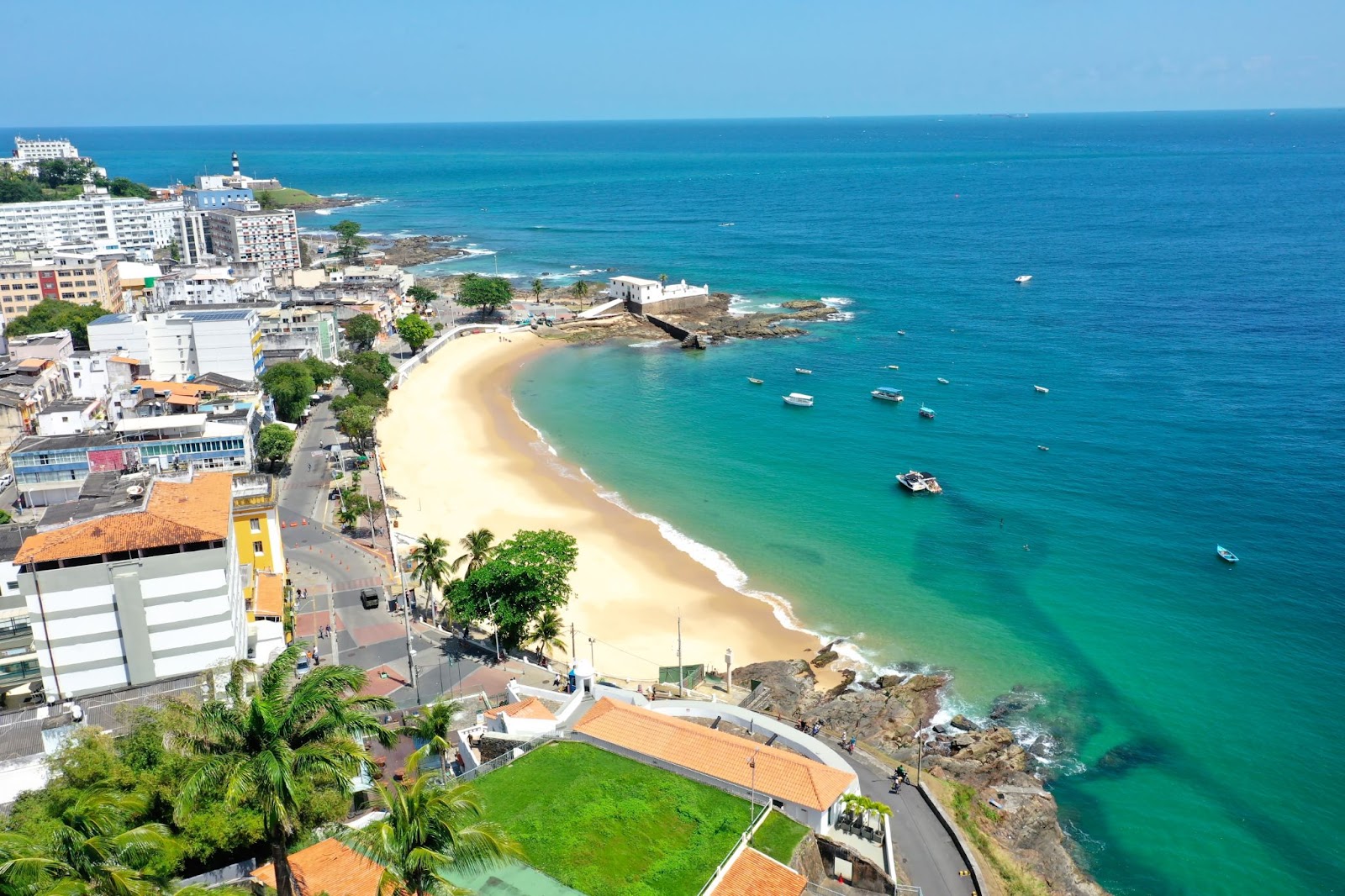 Vista aérea do litoral de Salvador, lar do Festival de Verão Salvador. O mar de cor azul intensa se encontra com uma faixa de areia clara. Depois dela, aparecem uma grande avenida e algumas construções baixas