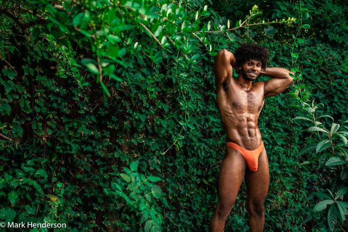 Tony Genius standing outside against a wall of shrubs wearing an orange thong with his hands behind his head