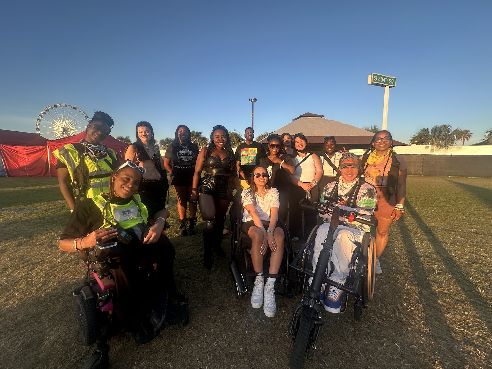 A group of BIPOC individuals participating in the Accessible+ Program poses for a photo on the grass field at Coachella. They are all smiling brightly and having fun.
