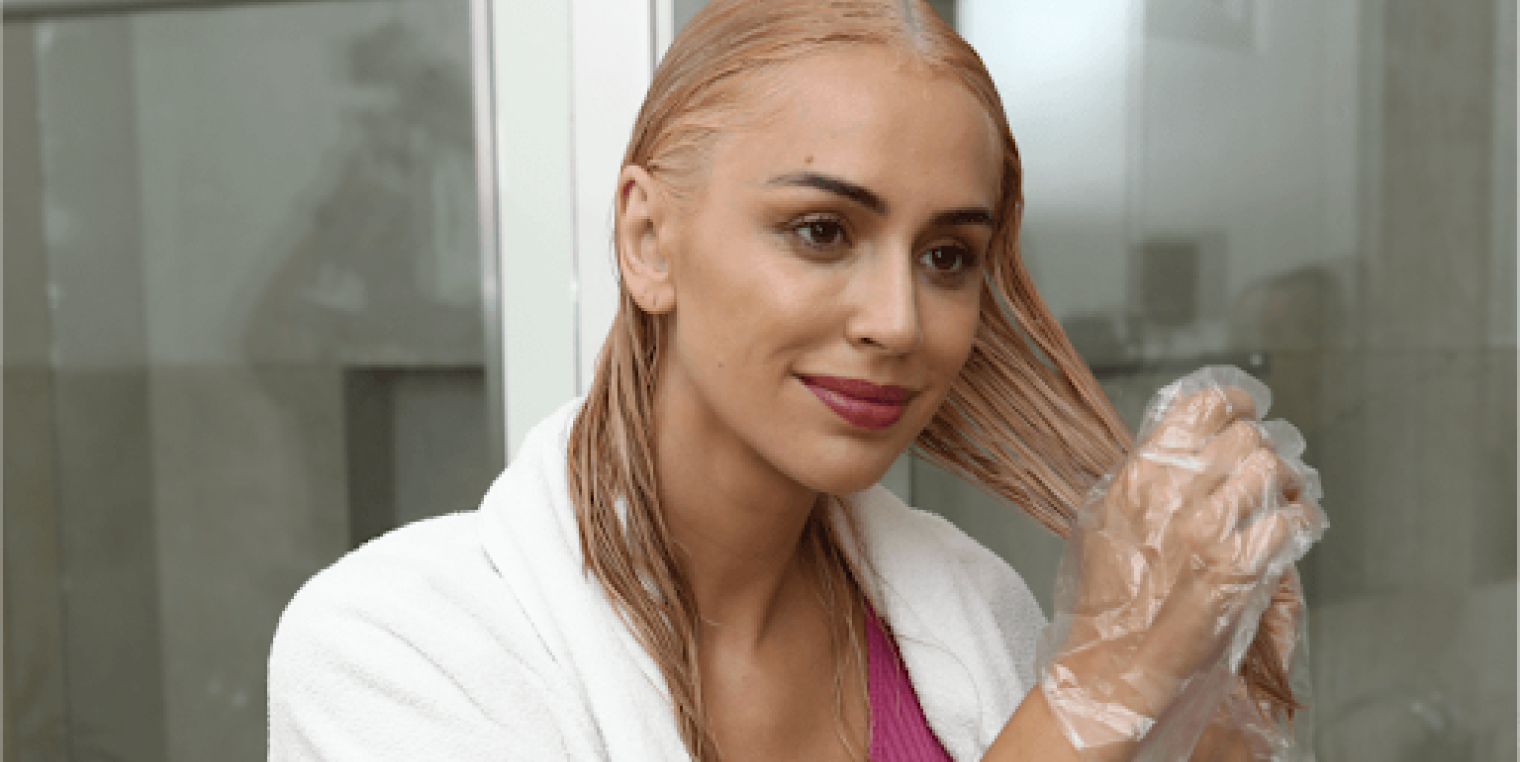 Imagem de uma mulher branca loira, com uma toalha no ombro, manuseando o cabelo com uma luva. Foto ilustrativa do processo de como pintar o cabelo em casa.