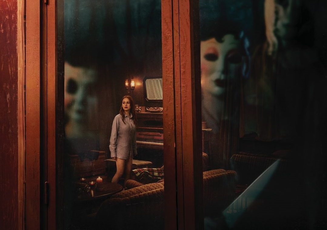 Una mujer con blusa celeste dentro de una casa, está siendo observada por dos personas con máscaras desde afuera de una ventana, con sus reflejos bien marcados en ella