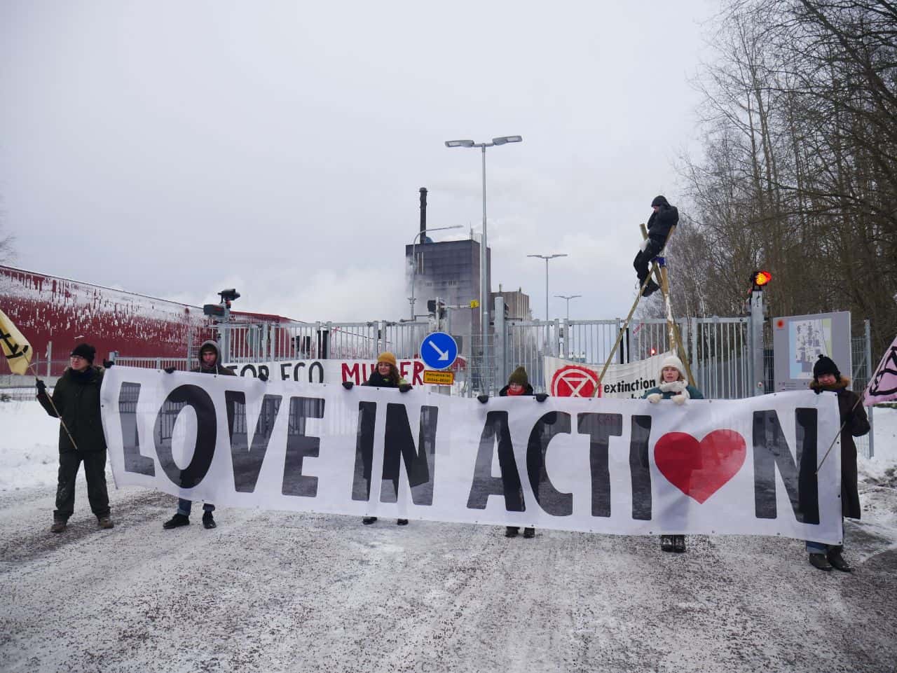 Rebels hold a banner that says Love In Action on a snowy road into a pulp mill.