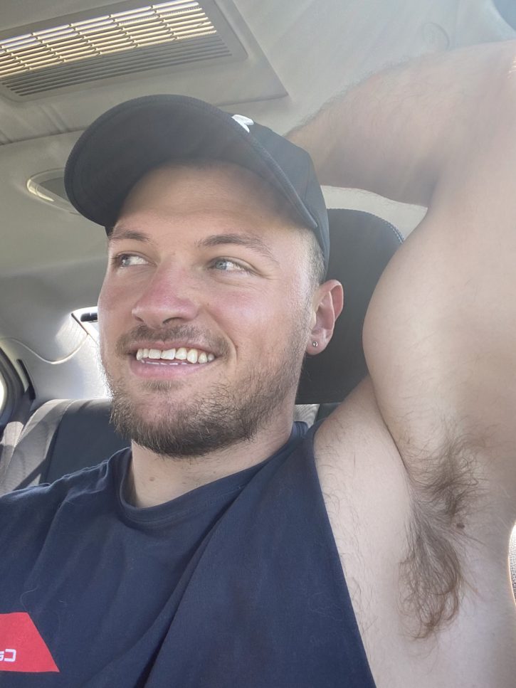 Max Small wearing a baseball hat and black tank top showing off his hairy arm pits while smiling in the car