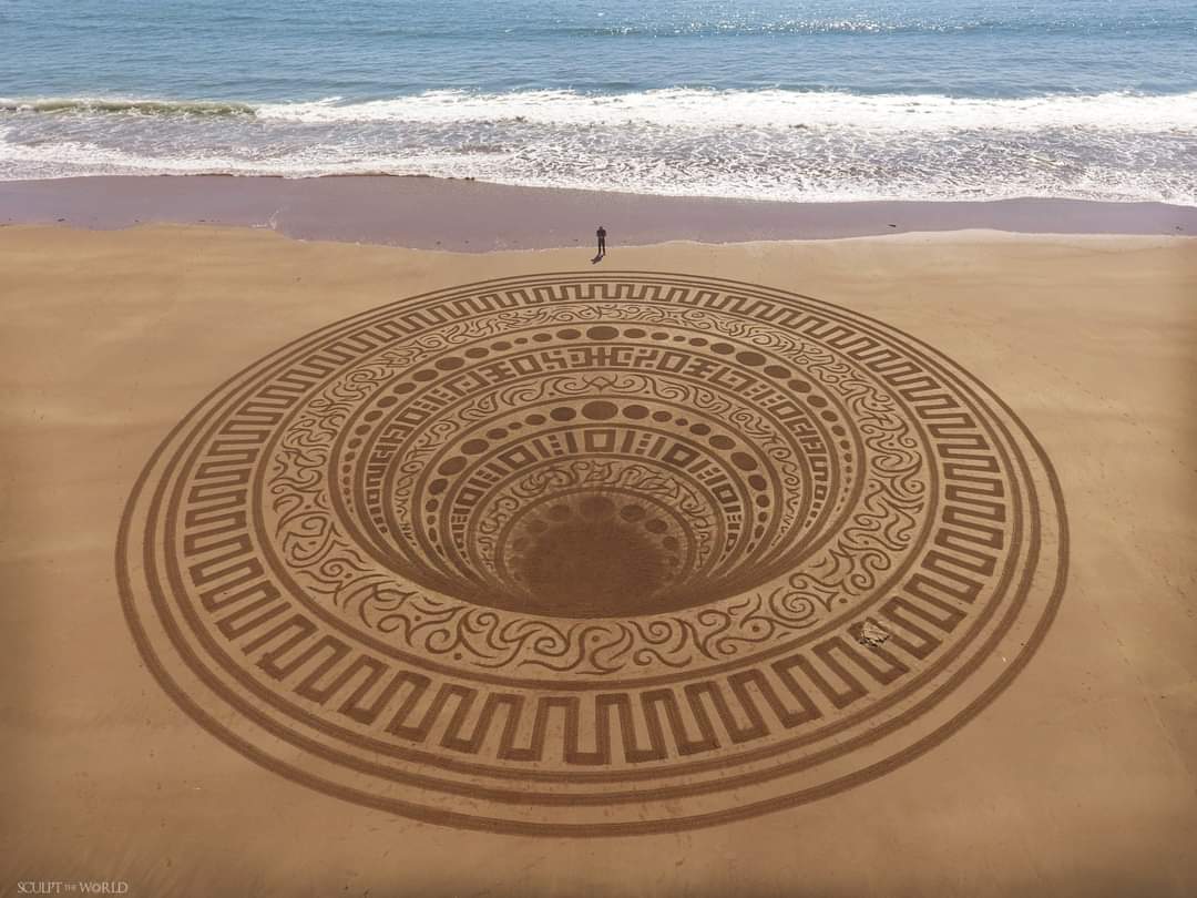 Photo of a large sand sculpture at the beach with the artist, Jon Foreman, standing above it.