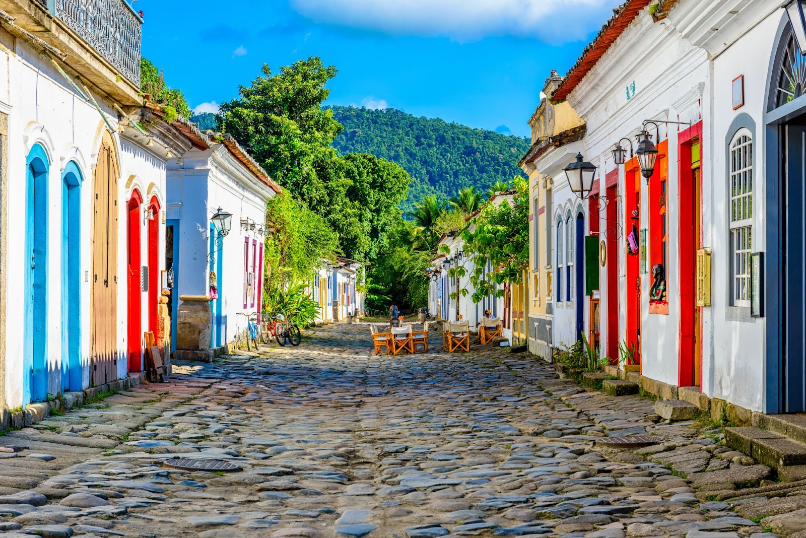 Rua do centro histórico de Paraty, com chão de paralelepípedo e casas coloniais brancas com detalhes em cores vivas como vermelho e azul. Ao horizonte, o verde da vegetação e da montanha fazem contraste com as cores
