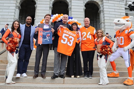 Broncos President Damani Leech, Denver Mayor Mike Johnston, Randy Gradishar, his wife beth, Governor Polis, Broncos Cheerleaders, and Miles the mascot post on the steps while holding a Gradishar jersey.