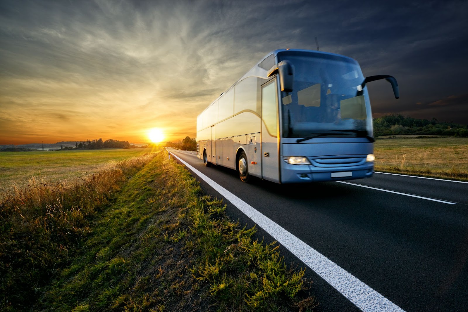 Ônibus de viagem em estrada vazia. Ao fundo, no horizonte atrás do ônibus, o sol está se pondo e iluminando toda a imagem com um tom amarelado.